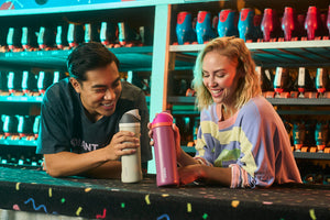 Two friends showing their grey and pink 24oz FreeSip water bottles to each other at a rollerskating center.